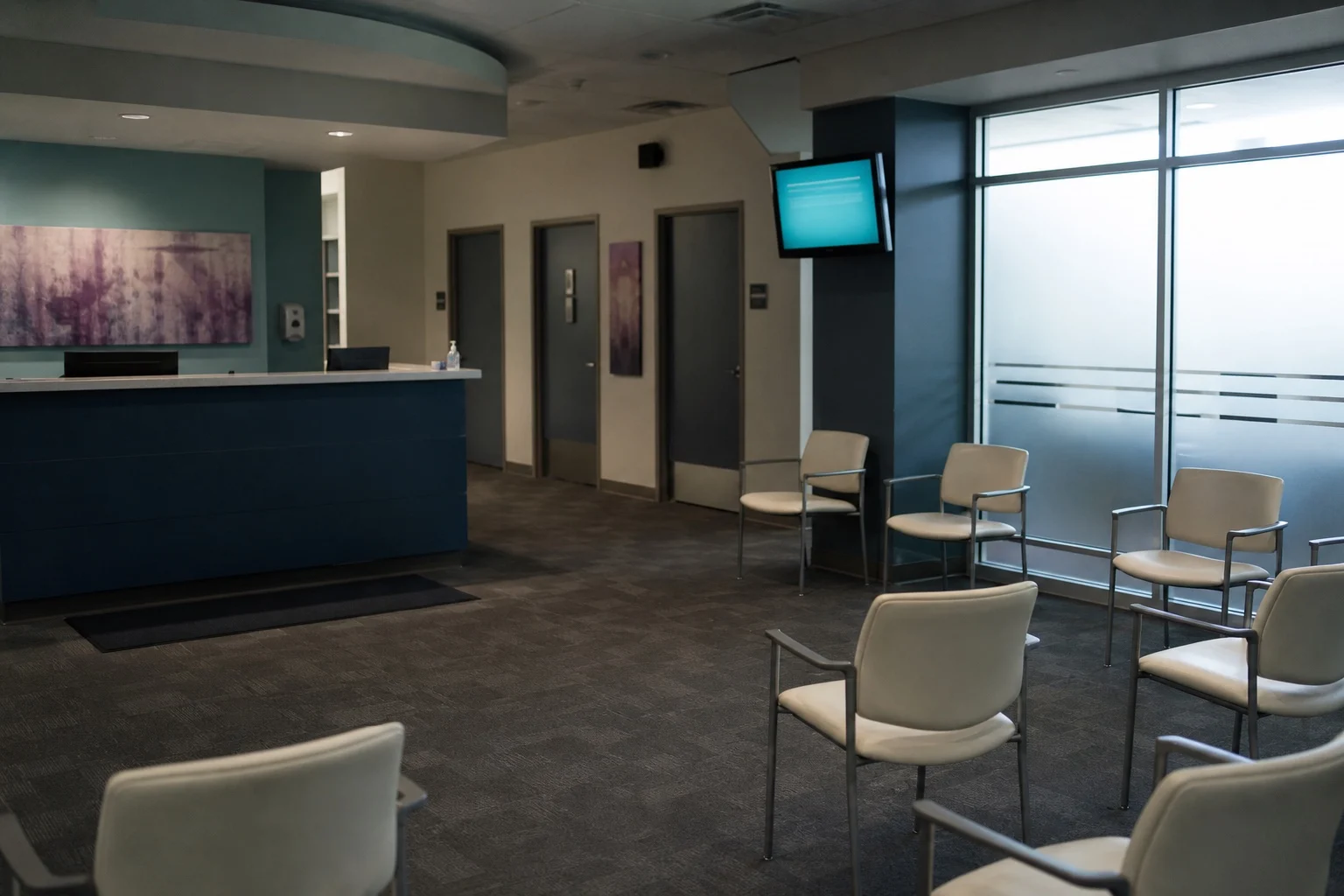 Empty medical clinic waiting room with spaced chairs and a quiet reception desk.